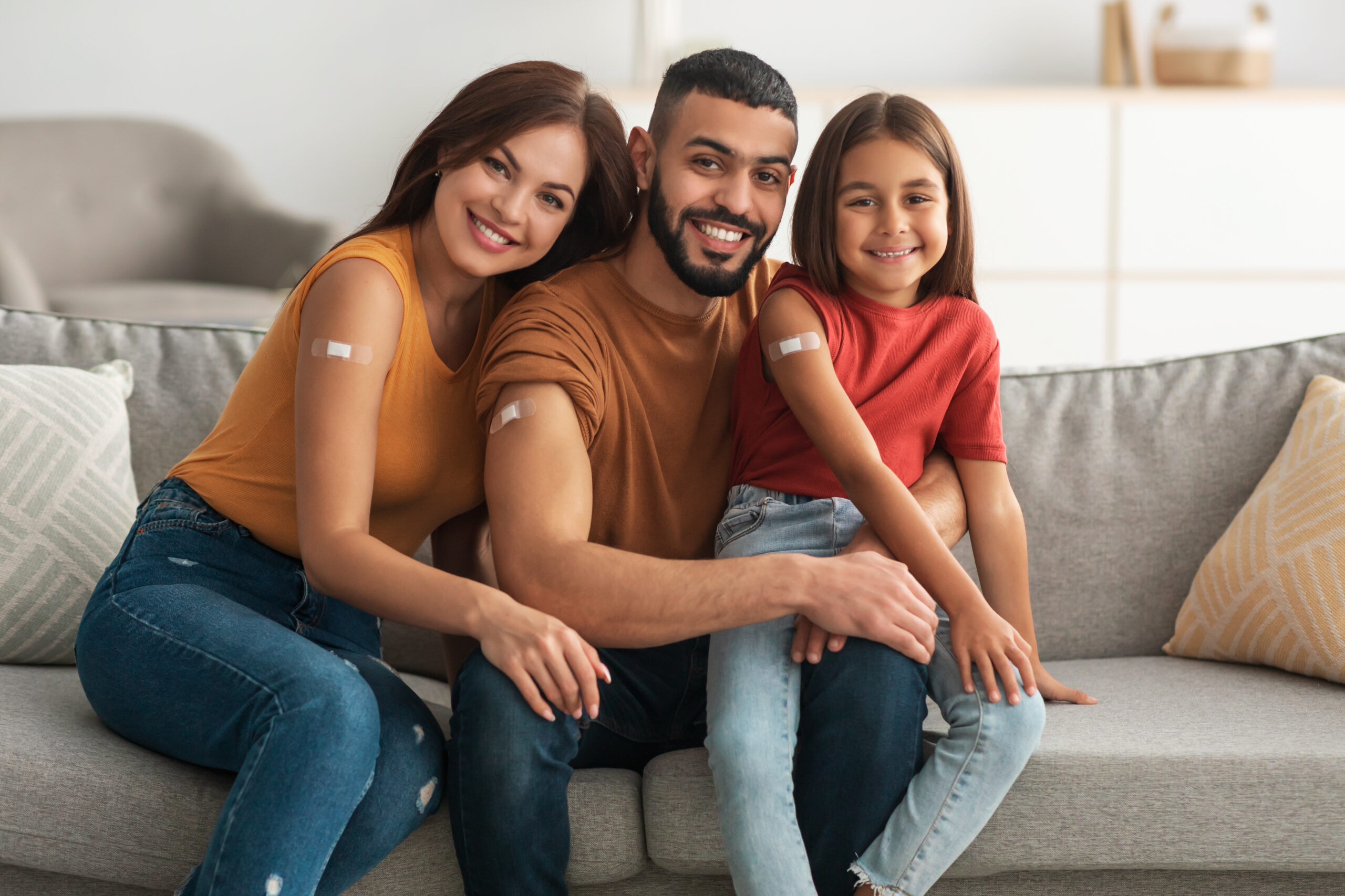 Coronavirus Vaccination Advert. Happy Vaccinated Family Of Three People Showing Arm With Sticking Plaster After Covid-19 Vaccine Vax Injection Posing Sitting On Couch In Living Room, Smiling To Camera