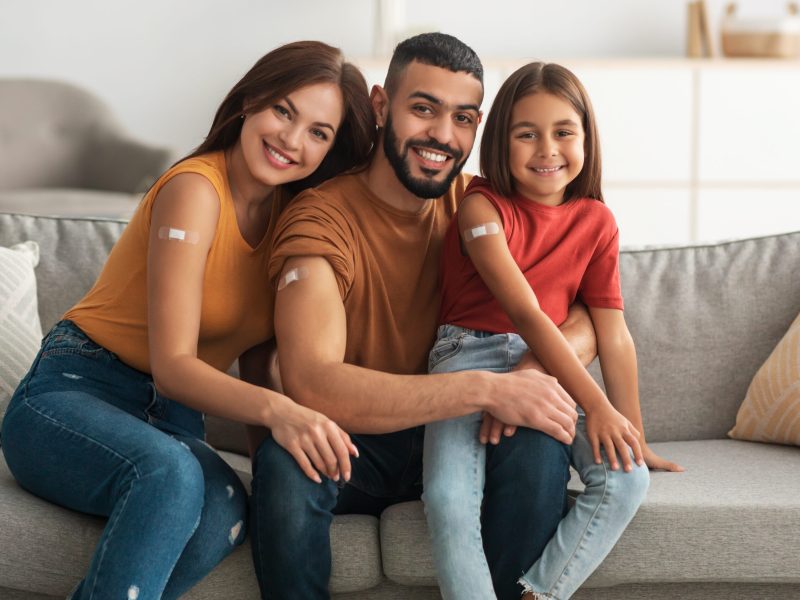 Coronavirus Vaccination Advert. Happy Vaccinated Family Of Three People Showing Arm With Sticking Plaster After Covid-19 Vaccine Vax Injection Posing Sitting On Couch In Living Room, Smiling To Camera